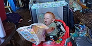 Caleb is registered to the contest to win money with this photo: baby, walker, flyer, living_room, clutter, bookshelf, book, toy, diapers, stroller, table, chair, carpet, plastic_container, camera, door, stuffed_animal, household_items, baby_supplies, person