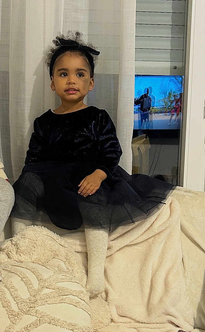 Djennah a rejoint le concours — aidez-le/la à gagner de superbes lots ! child, girl, black_dress, headband, couch, blanket, indoors, curtain, television, screen, window, sitting, portrait, cozy, smiling, person, furniture, home, living_room, curtains