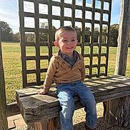 Roc is registered to the contest to win money with this photo: child, boy, smile, outdoor, bench, wood, jeans, sweater, boots, sunny, daylight, grass, field, blue_sky, happy, portrait, casual_clothing, nature, young, sitting