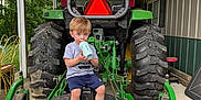Roc is registered to the contest to win money with this photo: toddler, child, bottle, tractor, farm_machinery, green, outdoor, building, shed, tires, clothing, footwear, curious, sitting, person, wood, metal, nature, leafy, caution_sign
