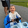 baby, child, smiling, happy, outdoor, push_car, toy_vehicle, steering_wheel, park, path, grass, trees, adult, parent, hands, white_outfit, blue_vehicle, safety_harness, portrait, family_outing