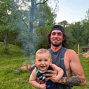 Emmett is registered to the contest to win money with this photo: adult, baby, baby_carrier, baseball_cap, campfire, child, family, father, grass, nature, outdoor, parent, portrait, sleeveless_shirt, smiling, smoke, summer, tattoo, trees, woodland