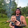 adult, baby, baby_carrier, baseball_cap, campfire, child, family, father, grass, nature, outdoor, parent, portrait, sleeveless_shirt, smiling, smoke, summer, tattoo, trees, woodland