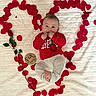 baby, infant, child, smiling, portrait, lying_down, red_clothing, white_blanket, rose_petals, heart_shape, rose, flower, milestone_photo, six_months, wooden_sign, bed, indoor, celebration, adorable, family_photo