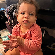 Angel is registered to the contest to win money with this photo: toddler, child, baby, pink_onesie, indoor, carpet, clapping_hands, curly_hair, face, expression, living_room, couch, diaper, skin, person, seated, smudge, home, casual, young_child