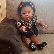 Delanie is registered to the contest to win money with this photo: baby, child, smiling, sitting, couch, curly_hair, bows, leopard_print, black_socks, indoor, cute, happy, person, infant, portrait, home, furniture, clothing, feet, expression