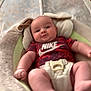 baby, infant, onesie, nike, baby_seat, face, legs, arms, indoor, soft_toys, comfort, cute, child, young, clothing, blurred_background, expression, skin, head, relaxing