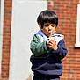 child, boy, dandelion, flower, outdoor, brick_wall, hoodie, curious, nature, blowing, seed, casual_clothing, daylight, young, expression, playful, plant, person, standing, puff