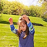 child, girl, outdoor, grass, sunlight, long_hair, blue_shirt, patterned_clothing, nature, trees, spring, greenery, smiling, playful, casual, daylight, happy, person, young, cute