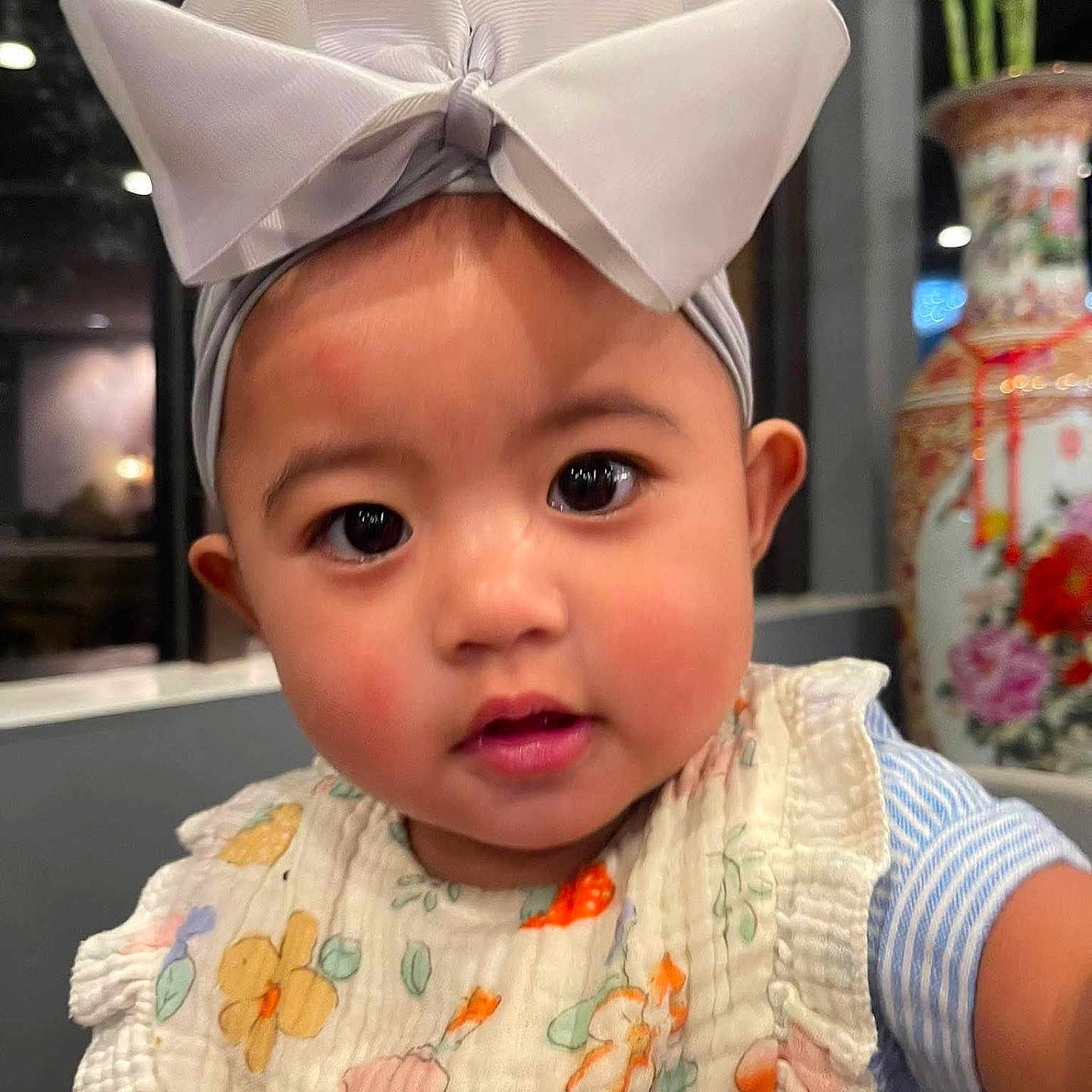 Kenzley Nery joined the competition — help win amazing prizes! baby, bib, bow, child, closeup, clothing, curious, cute, decor, expression, face, floral_pattern, headband, indoor, infant, person, plant, portrait, soft_lighting, vase