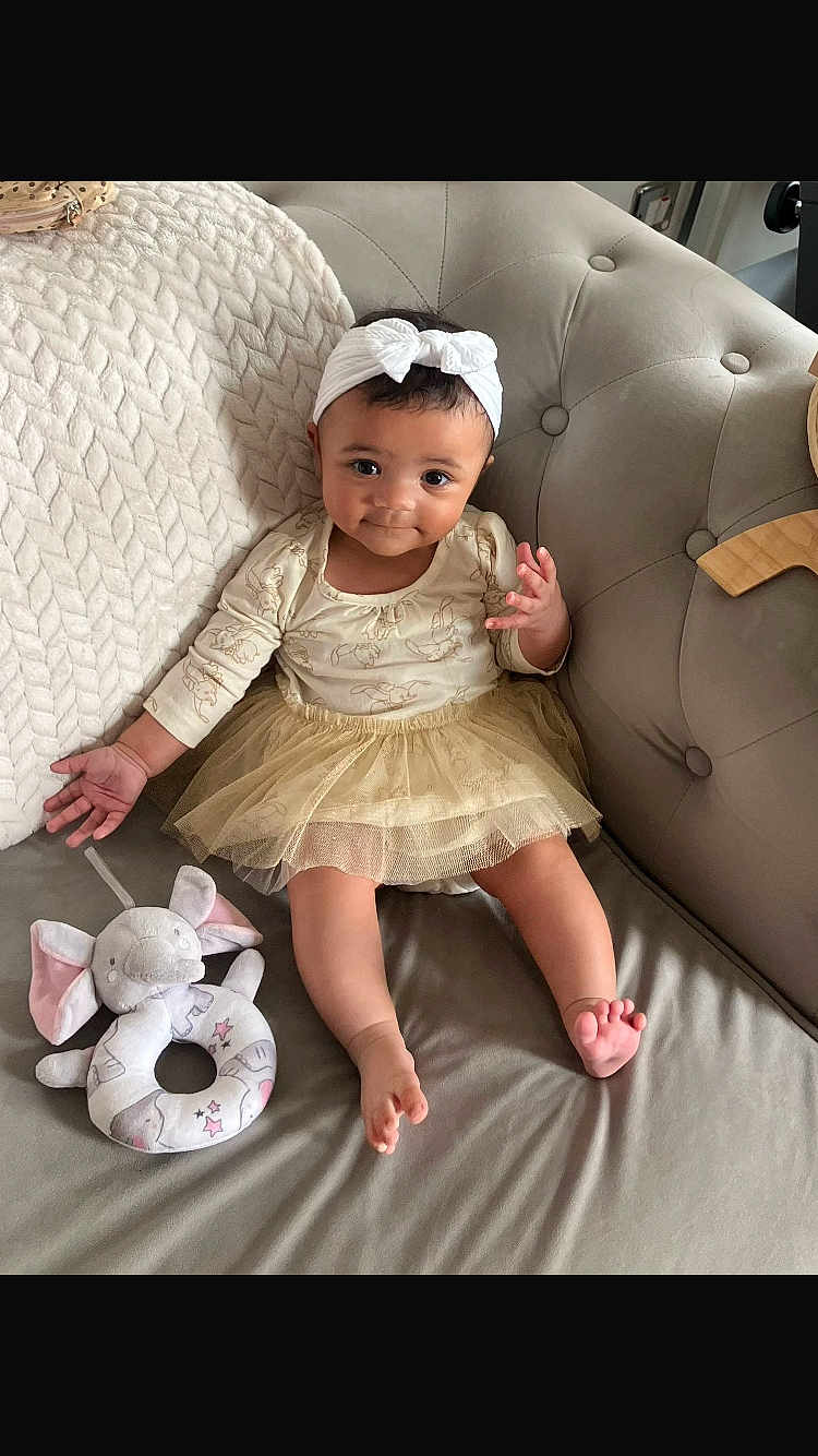 Talia joined the competition — help win amazing prizes! baby, child, headband, bow, dress, tulle_skirt, couch, plush_toy, elephant, knit_blanket, infant, smiling, indoor, cute, person, fabric, foot, hand, seated, home
