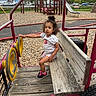 toddler, child, playground, bench, wood, toy_vehicle, slide, woodchips, outdoor, park, fence, trees, sky, clouds, car, parking_lot, clothing, hair_bun, sneakers, curly_hair