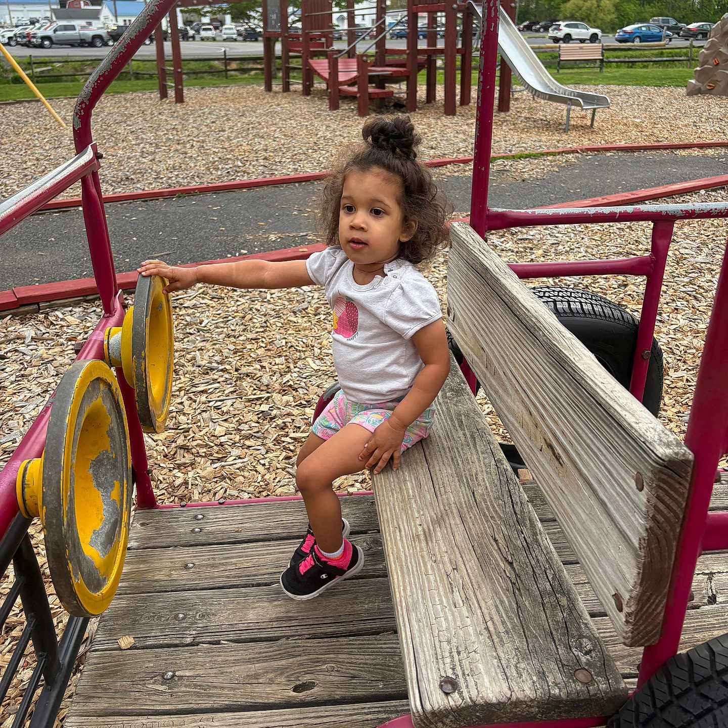 Taylor joined the competition — help win amazing prizes! toddler, child, playground, bench, wood, toy_vehicle, slide, woodchips, outdoor, park, fence, trees, sky, clouds, car, parking_lot, clothing, hair_bun, sneakers, curly_hair