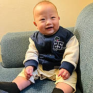 Aaron joined the competition — help win amazing prizes! baby, smiling, sitting, couch, blue_couch, jacket, dinosaur_patch, number_25, shorts, black_socks, indoor, child, happy, portrait, face, person, infant, cute, wall, cozy