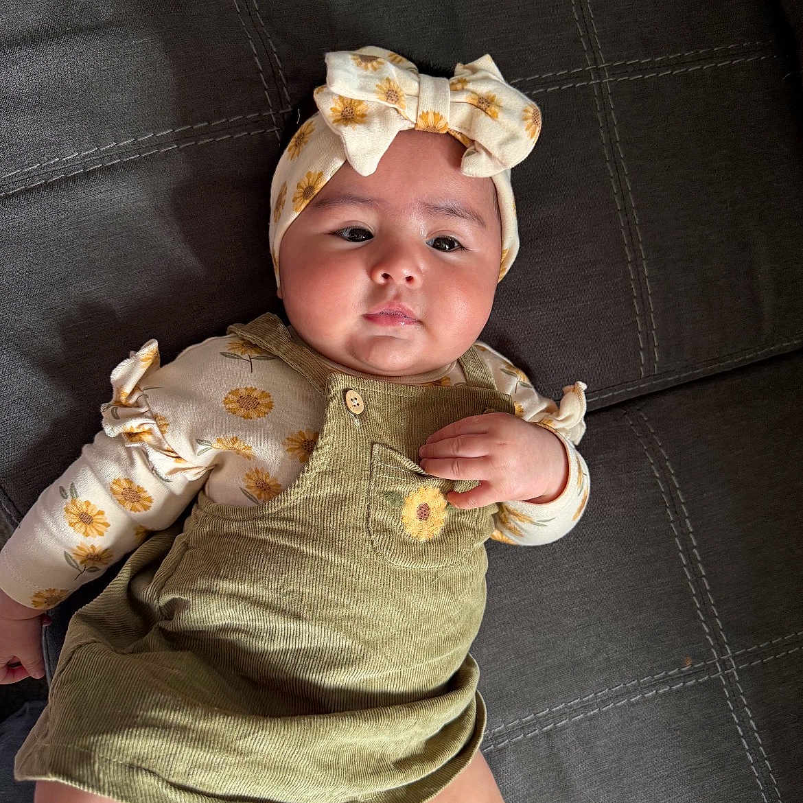 Jailani is registered to the contest to win money with this photo: baby, child, infant, portrait, lying_down, indoors, headband, bow, floral_pattern, sunflowers, overalls, dress, cushion, sofa, textile, smiling, face, hands, fashion, photography