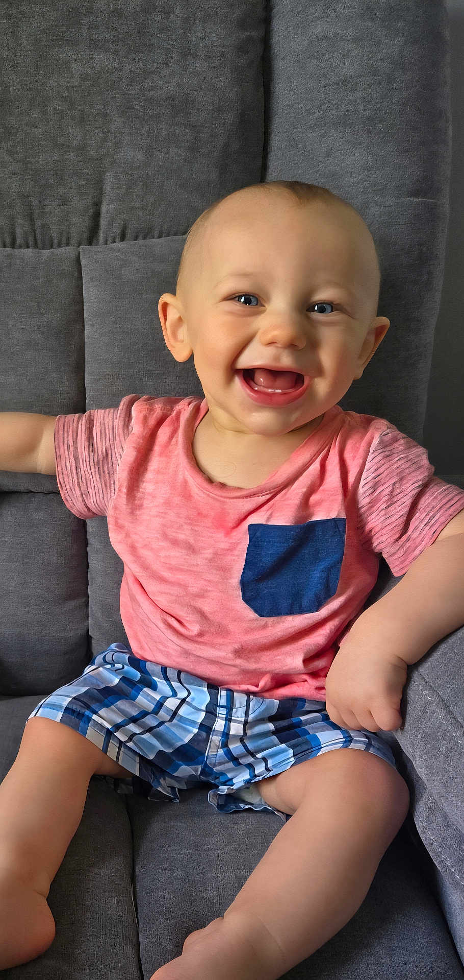 Connor joined the competition — help win amazing prizes! baby, child, smiling, happy, sitting, couch, clothing, pink_shirt, blue_pocket, plaid_shorts, face, person, indoor, cute, infant, legs, arm, furniture, blue_eyes, skin