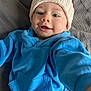 baby, child, blue_hoodie, knit_hat, smiling, face, person, infant, happy, cute, apparel, portrait, laying_down, young_child, indoors, soft_texture, gray_background, clothing, close_up, headwear