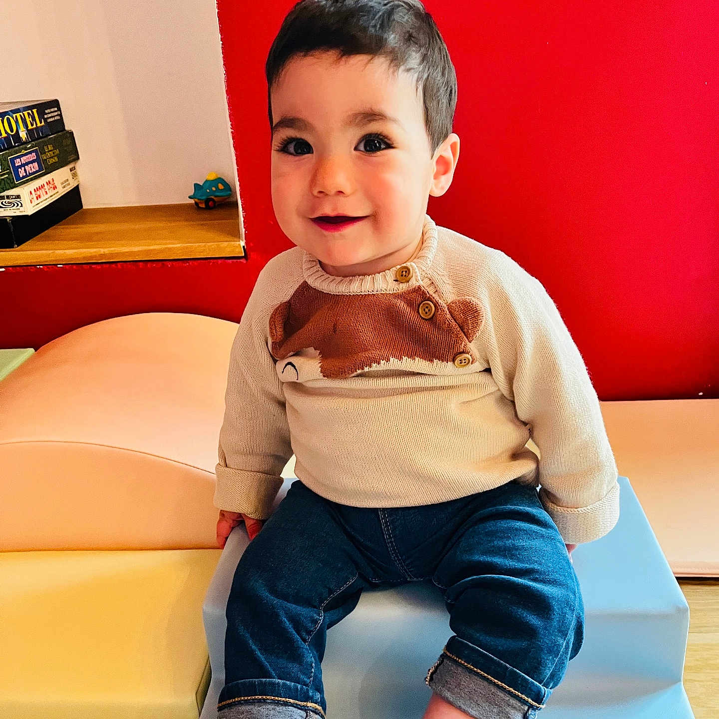 Jules participe au concours pour gagner de l'argent avec cette photo : casual_clothing, child, cute, face, furniture, happy, indoor, jeans, person, playroom, portrait, red_wall, shoes, sitting, smiling, sweater, toddler, toy, wooden_floor, young_child