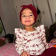 Reign is registered to the contest to win money with this photo: bed, bracelet, child, cozy, cute, face, happy, headwrap, home, indoors, jewelry, maroon, pants, person, pink_bedspread, red_bows, sitting, smiling, toddler, white_top
