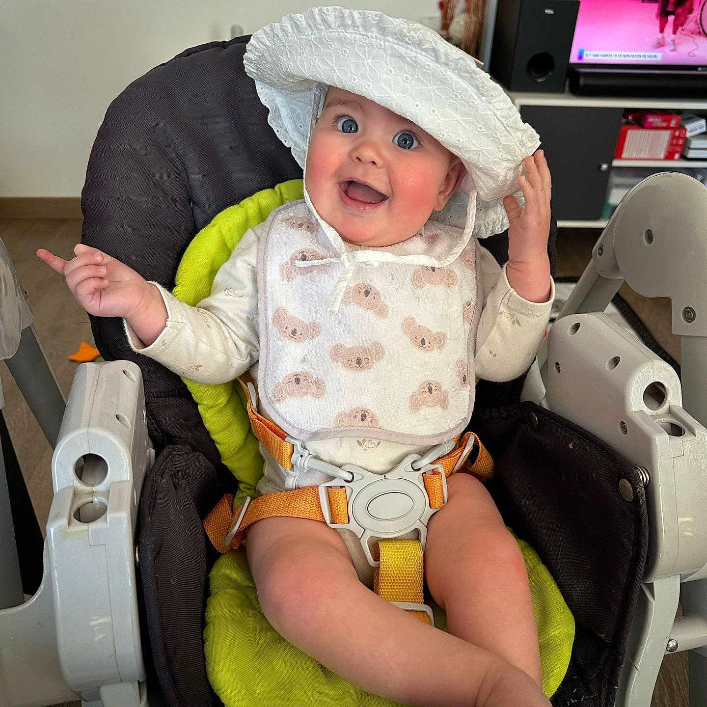 Elynna a rejoint le concours — aidez-le/la à gagner de superbes lots ! baby, bib, child, clothing, expression, face, floor, foot, furniture, hand, happy, hat, high_chair, indoor, person, pointing, screen, smile, television, wood