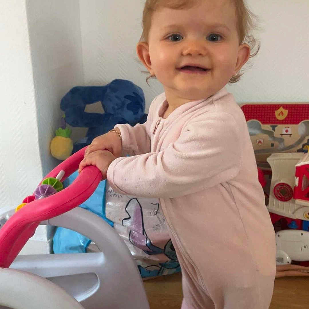 Elyana participe au concours pour gagner de l'argent avec cette photo : baby, background, child, cute, face, floor, footwear, hair, hand, happy, indoor, person, pink_onesie, playroom, smiling, standing, toddler, toy, toy_car, walker