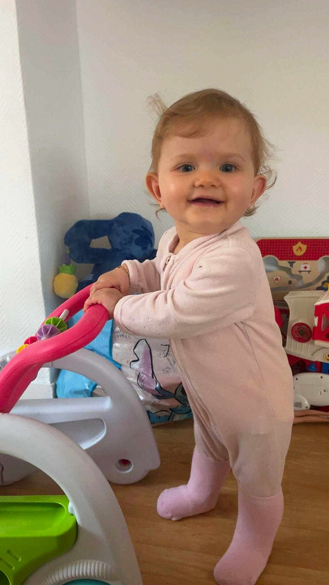 Elyana participe au concours pour gagner de l'argent avec cette photo : toddler, child, smiling, standing, pink_onesie, toy, walker, indoor, floor, face, person, baby, playroom, cute, happy, footwear, hair, hand, toy_car, background
