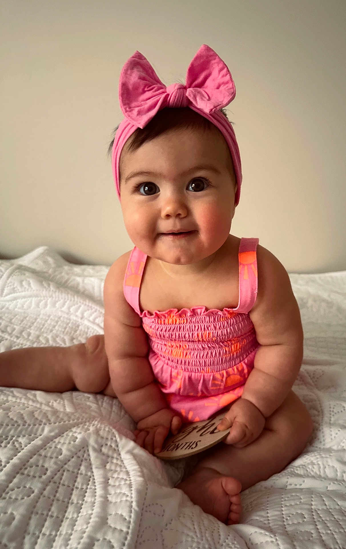 Kora Belle is registered to the contest to win money with this photo: baby, child, pink_bow, headband, dress, sitting, blanket, quilt, milestone_card, cute, chubby_cheeks, indoor, portrait, smiling, infant, hands, feet, curious_eyes, soft_lighting, skin