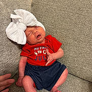 Mary is registered to the contest to win money with this photo: baby, newborn, infant, sleeping, bow, headwear, red_shirt, blue_shorts, couch, fabric, hand, peaceful, indoors, skin, person, clothing, textile, seated, small, cute