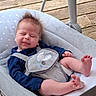 baby, infant, child, baby_seat, cushion, wooden_deck, blue_clothing, gray_harness, sheep_design, closed_eyes, tongue_out, bare_feet, spiky_hair, playful_expression, outdoor, daylight, cute, relaxed, person, infant_care