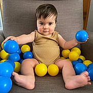 Nolan participe au concours pour gagner de l'argent avec cette photo : toddler, child, baby, armchair, plastic_balls, blue_ball, yellow_ball, clothing, onesie, indoor, furniture, play, sitting, barefoot, cute, expression, person, face, hair, toy