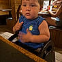 toddler, child, blue_tank_top, pineapple_logo, high_chair, wooden_table, restaurant, indoor, dining, person, face, arm, furniture, seat, food, casual_clothing, table, background_people, light, tile_floor