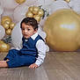 toddler, child, boy, blue_clothing, vest, polka_dot_shirt, barefoot, sitting, wooden_floor, balloons, gold_balloons, white_balloons, party_decoration, indoor, portrait, curious_expression, young_child, celebration, decor, flooring