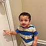 toddler, child, boy, striped_shirt, blue_shirt, white_shirt, door, door_knob, hand, carpet, star_pattern, indoor, wall, beige_wall, person, standing, young_child, curly_hair, smiling, cute