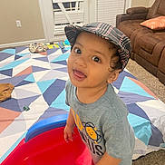Yuvaan is registered to the contest to win money with this photo: toddler, child, indoor, plaid_cap, grey_shirt, toy, red_toy, blue_toy, carpet, blanket, geometric_pattern, brown_recliner, living_room, curious, playful, smiling, wide_eyes, person, furniture, home