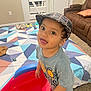 toddler, child, indoor, plaid_cap, grey_shirt, toy, red_toy, blue_toy, carpet, blanket, geometric_pattern, brown_recliner, living_room, curious, playful, smiling, wide_eyes, person, furniture, home