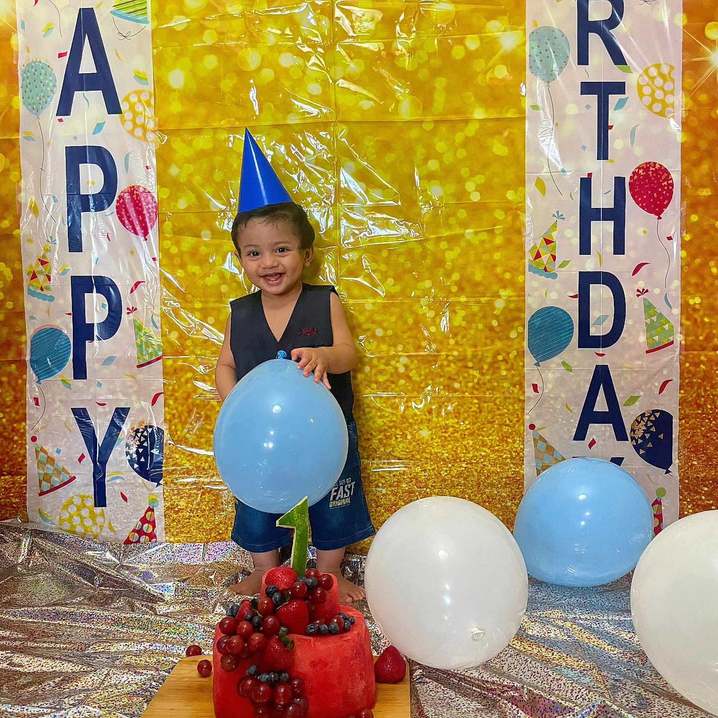 Yuvaan is registered to the contest to win money with this photo: balloon, birthday, blue_balloon, blueberries, celebration, child, colorful_backdrop, confetti, festive, first_birthday, golden_background, grapes, indoor, party_banner, party_hat, silver_floor, smiling_child, toddler, watermelon_cake, white_balloon