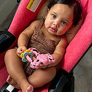 Khloe is registered to the contest to win money with this photo: baby, child, stroller, toy, pink, curly_hair, cute, infant, playful, indoors, face, person, holding, seat, clothing, skin, foot, hand, expression, comfort
