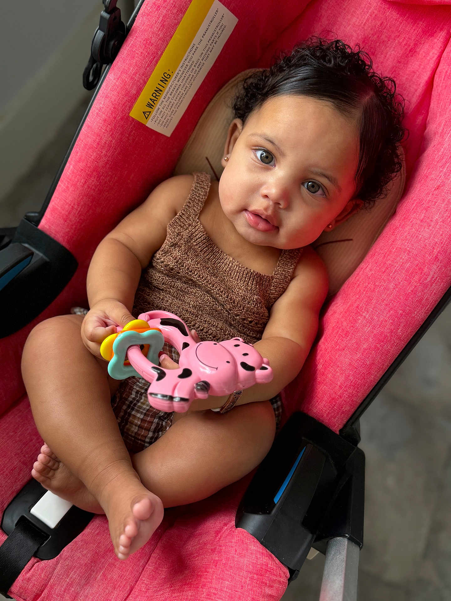 Khloe is registered to the contest to win money with this photo: baby, child, stroller, toy, pink, curly_hair, cute, infant, playful, indoors, face, person, holding, seat, clothing, skin, foot, hand, expression, comfort