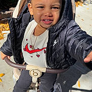 Khloe is registered to the contest to win money with this photo: child, girl, toddler, portrait, smiling, outdoor, stroller, sitting, red_bows, braids, jacket, shirt, necklace, earrings, shoes, sunlight, sidewalk, garden, leaves, photography