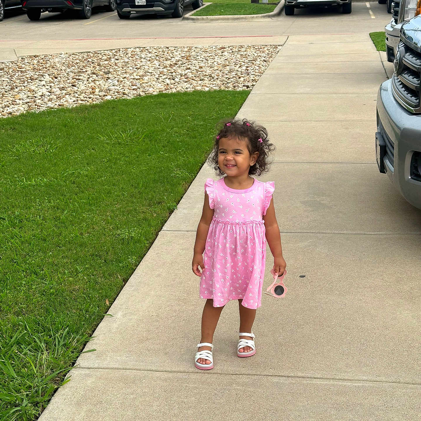 Isabella joined the competition — help win amazing prizes! cars, casual, child, concrete, curly_hair, cute, daytime, girl, grass, happy, outdoor, parking_lot, person, pink_dress, playful, sandals, sidewalk, smiling, sunglasses, urban