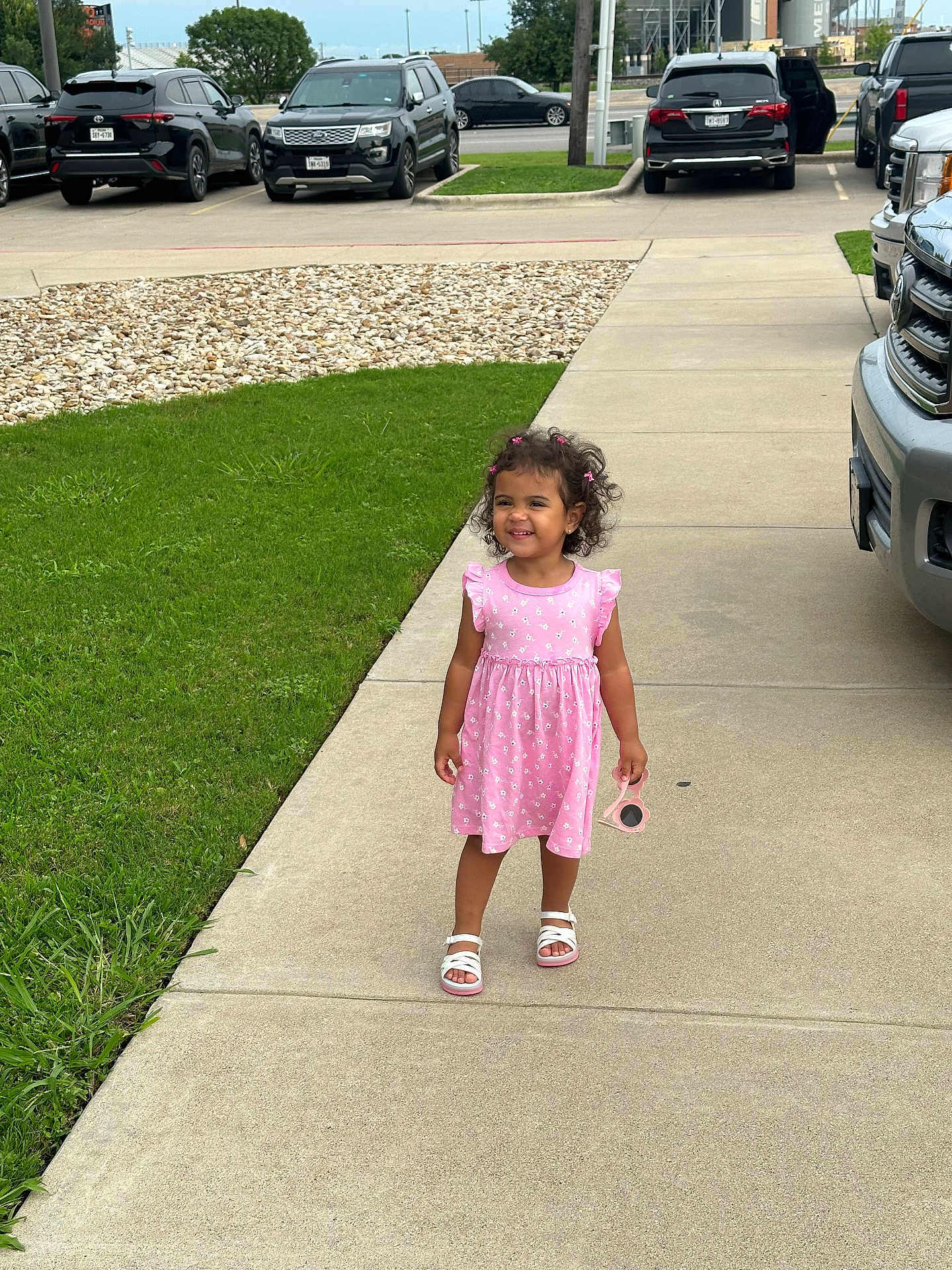 Isabella joined the competition — help win amazing prizes! child, girl, pink_dress, sunglasses, sidewalk, grass, cars, parking_lot, smiling, curly_hair, sandals, outdoor, daytime, happy, person, concrete, urban, casual, cute, playful