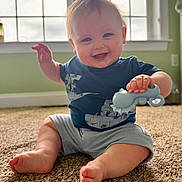 Grayson is registered to the contest to win money with this photo: baby, smiling, toy, carpet, indoor, child, happy, sitting, blue_shirt, shorts, window, green_wall, cute, infant, hand, foot, playing, person, portrait, home
