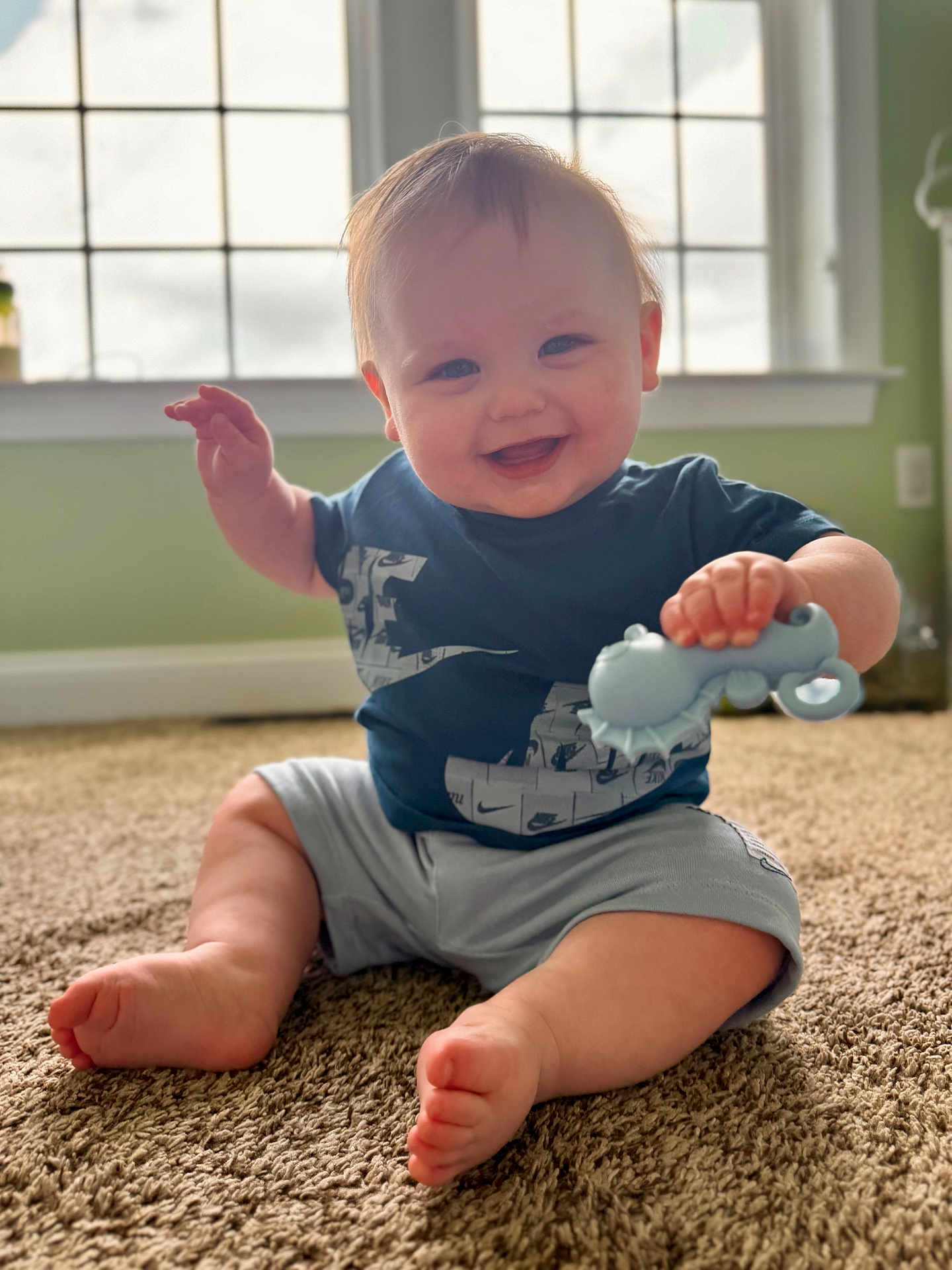 Grayson is registered to the contest to win money with this photo: baby, smiling, toy, carpet, indoor, child, happy, sitting, blue_shirt, shorts, window, green_wall, cute, infant, hand, foot, playing, person, portrait, home