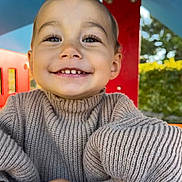 Yüna participe au concours pour gagner de l'argent avec cette photo : child, toddler, smile, face, sweater, knitwear, beige, outdoor, playground, red_structure, happy, portrait, person, nature, greenery, daylight, cute, young_child, closeup, hands