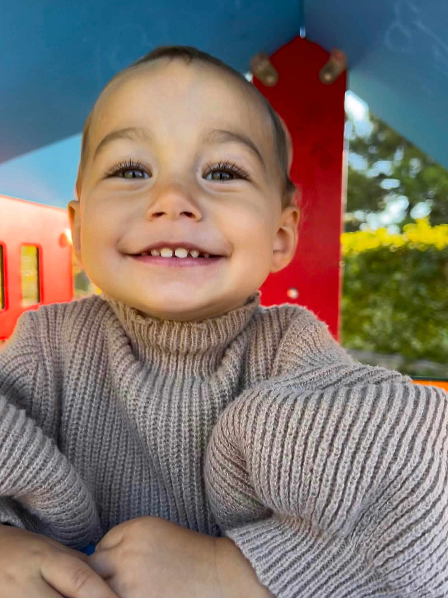 Yüna participe au concours pour gagner de l'argent avec cette photo : child, toddler, smile, face, sweater, knitwear, beige, outdoor, playground, red_structure, happy, portrait, person, nature, greenery, daylight, cute, young_child, closeup, hands