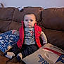 baby, child, red_vest, gray_shirt, socks, brown_couch, pillow, blue_pillow, beige_pillow, indoor, seated, person, cute, home, furniture, portrait, expressionless, comfortable, casual_clothing, young_child