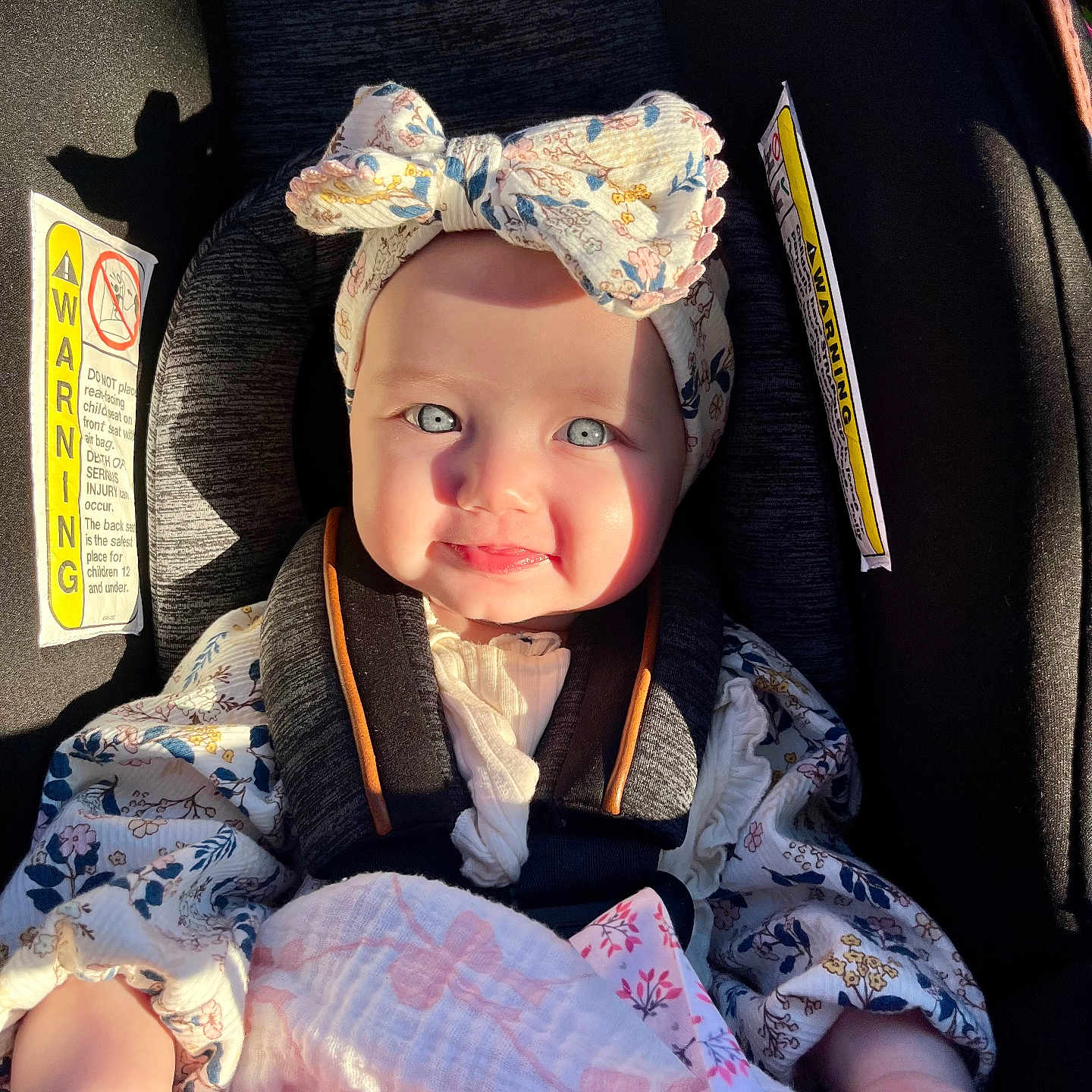 Melanie is registered to the contest to win money with this photo: baby, child, car_seat, headband, bow, blanket, smile, face, sitting, sunlight, shadow, clothing, infant, safety_harness, cute, portrait, indoor, person, happy, flower_pattern