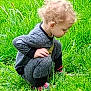 child, toddler, grass, greenery, outdoor, curly_hair, gray_clothing, nature, crouching, side_view, young_child, casual_wear, exploring, field, daylight, shoes, focused, person, small_child, curious