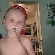 Austin is registered to the contest to win money with this photo: toddler, child, pacifier, red_bandana, shirtless, indoor, baby_gate, window, doorway, household, person, young_child, face, expressionless, blond_hair, toy, room, lighting, curtains, wall