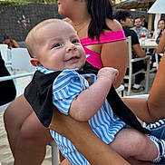 Liam a rejoint le concours — aidez-le/la à gagner de superbes lots ! baby, smile, striped_clothing, person, adult_hand, outdoor, people, table, chair, casual, summer, happy, infant, blue_clothing, black_clothing, shorts, hairstyle, tattoo, glasses, social_gathering